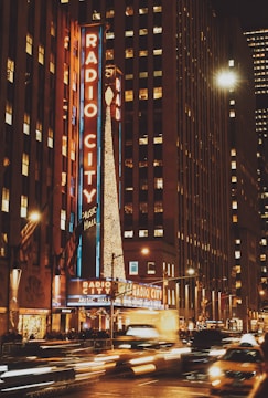 Bright neon lights illuminate a bustling city street. The iconic marquee of Radio City Music Hall glows in vibrant colors, while light trails from passing cars create a dynamic feel. Tall buildings surround the area, adding to the urban atmosphere.