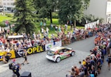A rally car speeds along a cobblestone street lined with spectators who are eagerly watching the event. The crowd is behind barricades, and many people are capturing the moment with cameras and phones. Banners for sponsors like Opel and other advertisements are visible. The setting appears to be a sunny day with people dressed in summer attire.