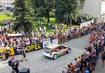 A rally car speeds along a cobblestone street lined with spectators who are eagerly watching the event. The crowd is behind barricades, and many people are capturing the moment with cameras and phones. Banners for sponsors like Opel and other advertisements are visible. The setting appears to be a sunny day with people dressed in summer attire.