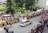 A rally car speeds along a cobblestone street lined with spectators who are eagerly watching the event. The crowd is behind barricades, and many people are capturing the moment with cameras and phones. Banners for sponsors like Opel and other advertisements are visible. The setting appears to be a sunny day with people dressed in summer attire.