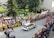A rally car speeds along a cobblestone street lined with spectators who are eagerly watching the event. The crowd is behind barricades, and many people are capturing the moment with cameras and phones. Banners for sponsors like Opel and other advertisements are visible. The setting appears to be a sunny day with people dressed in summer attire.