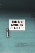A teal brick wall features a prominent white sign that reads 'This is a SMOKING AREA' in bold black letters. Below the text are graphic symbols of cigarettes indicating permitted smoking. An ashtray is mounted on the wall to the left of the sign, used to dispose of cigarette butts.