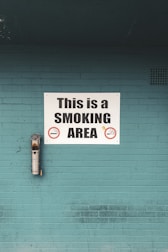 A teal brick wall features a prominent white sign that reads 'This is a SMOKING AREA' in bold black letters. Below the text are graphic symbols of cigarettes indicating permitted smoking. An ashtray is mounted on the wall to the left of the sign, used to dispose of cigarette butts.