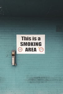 A teal brick wall features a prominent white sign that reads 'This is a SMOKING AREA' in bold black letters. Below the text are graphic symbols of cigarettes indicating permitted smoking. An ashtray is mounted on the wall to the left of the sign, used to dispose of cigarette butts.