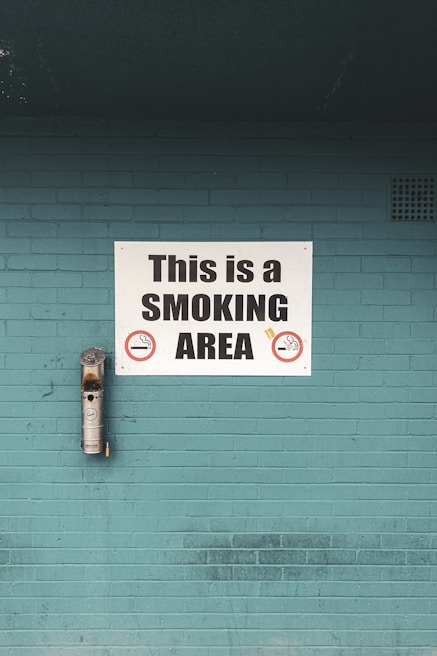 A teal brick wall features a prominent white sign that reads 'This is a SMOKING AREA' in bold black letters. Below the text are graphic symbols of cigarettes indicating permitted smoking. An ashtray is mounted on the wall to the left of the sign, used to dispose of cigarette butts.