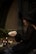A person is dressed in a dark, flowing gown, wearing a large, pointed hat, suggestive of a witch. They are holding an animal skull in their hands. The setting is dimly lit, possibly evening or in a shaded area, with a mysterious and mystical ambiance. Beside them are several black candles, some lit, and a few white pumpkins on the ground.