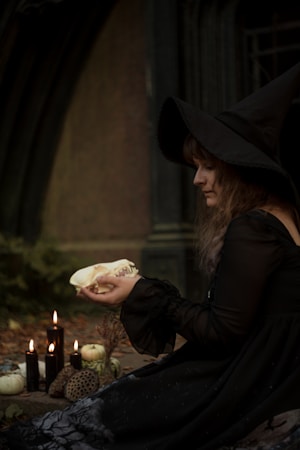 A person is dressed in a dark, flowing gown, wearing a large, pointed hat, suggestive of a witch. They are holding an animal skull in their hands. The setting is dimly lit, possibly evening or in a shaded area, with a mysterious and mystical ambiance. Beside them are several black candles, some lit, and a few white pumpkins on the ground.