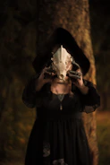 A mysterious figure wearing dark clothing holds up an animal skull in front of a tree in a dimly-lit forest setting. The person is partially obscured by the skull and a hood casts shadows over their face.