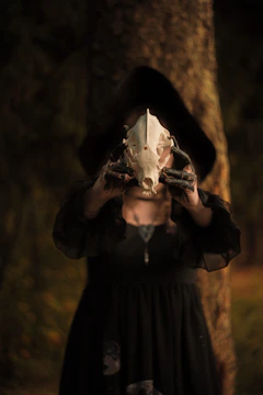 A mysterious figure wearing dark clothing holds up an animal skull in front of a tree in a dimly-lit forest setting. The person is partially obscured by the skull and a hood casts shadows over their face.