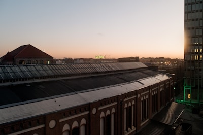 A cityscape at dusk featuring a large brick building with a series of windows and a sloped roof in the foreground. In the background, a skyline with various buildings is visible under a pale sunset sky. A prominent sign with the text 'METRO' is illuminated in the distance. The right side of the image includes a modern glass-paneled building.