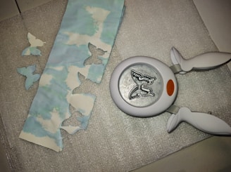 A butterfly-shaped paper punch is lying on a textured silver surface next to a strip of paper with cloud patterns. The paper has several butterfly-shaped cutouts, and a few of these cutouts are placed nearby on the surface.