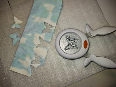 A butterfly-shaped paper punch is lying on a textured silver surface next to a strip of paper with cloud patterns. The paper has several butterfly-shaped cutouts, and a few of these cutouts are placed nearby on the surface.