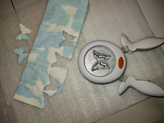 A butterfly-shaped paper punch is lying on a textured silver surface next to a strip of paper with cloud patterns. The paper has several butterfly-shaped cutouts, and a few of these cutouts are placed nearby on the surface.