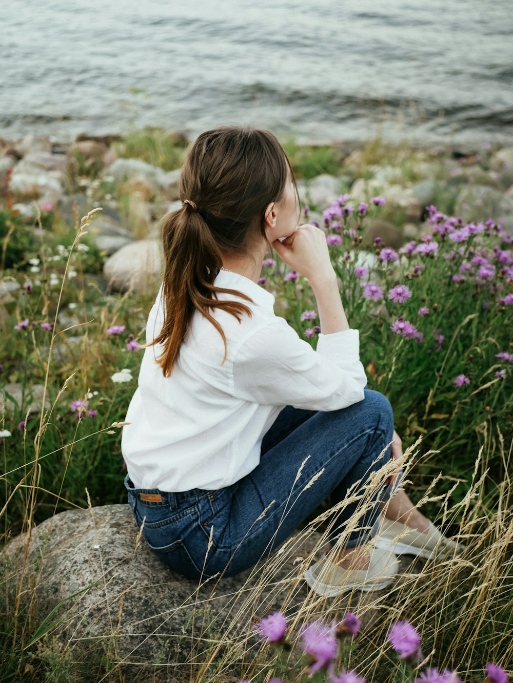 Donna in camicia bianca a maniche lunghe e jeans blu in denim seduta sulla roccia vicino a fiori viola