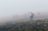 People are walking through a foggy landscape, with visibility reduced by the dense mist. The ground appears grassy, and individuals wear jackets and carry backpacks, suggesting an outdoor hike or journey.