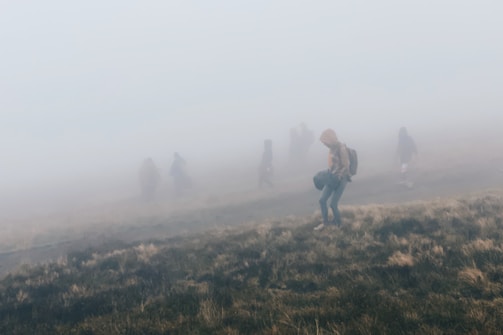 People are walking through a foggy landscape, with visibility reduced by the dense mist. The ground appears grassy, and individuals wear jackets and carry backpacks, suggesting an outdoor hike or journey.