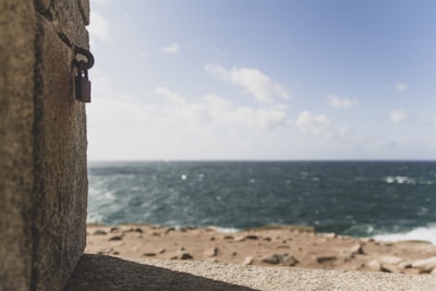 A padlock is attached to a stone wall in the foreground, with a clear view of the ocean stretching into the horizon under a blue sky filled with scattered clouds.