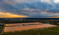A large agricultural greenhouse complex with transparent roofs is illuminated by artificial lighting. The complex is surrounded by greenery and situated near a cityscape visible in the distant background. The sky is overcast with dark clouds, and there is a faint orange and yellow glow near the horizon suggesting a sunset or sunrise.
