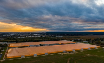 A large agricultural greenhouse complex with transparent roofs is illuminated by artificial lighting. The complex is surrounded by greenery and situated near a cityscape visible in the distant background. The sky is overcast with dark clouds, and there is a faint orange and yellow glow near the horizon suggesting a sunset or sunrise.