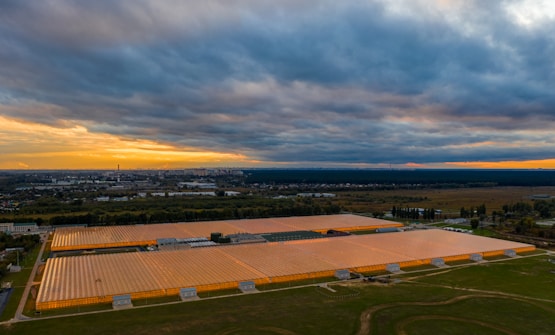 A large agricultural greenhouse complex with transparent roofs is illuminated by artificial lighting. The complex is surrounded by greenery and situated near a cityscape visible in the distant background. The sky is overcast with dark clouds, and there is a faint orange and yellow glow near the horizon suggesting a sunset or sunrise.