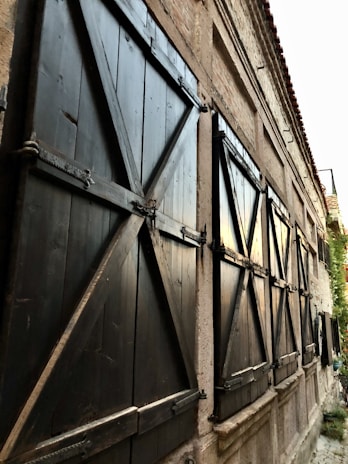 A row of large, closed wooden shutters with a lattice design, mounted on an old building with a textured stone facade. The shutters are painted black and secured with metal braces, and the setting appears to be a narrow alley with soft evening light reflecting off the windows.