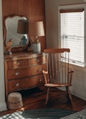 A cozy corner of a room features a wooden dresser with a mirror on top, a classic wooden chair, a table lamp with a beige shade, and a decorative bird figurine. The light from the window with blinds softly illuminates the wooden furniture and creates a warm atmosphere.