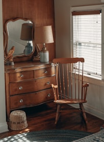 A cozy corner of a room features a wooden dresser with a mirror on top, a classic wooden chair, a table lamp with a beige shade, and a decorative bird figurine. The light from the window with blinds softly illuminates the wooden furniture and creates a warm atmosphere.
