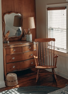 A cozy corner of a room features a wooden dresser with a mirror on top, a classic wooden chair, a table lamp with a beige shade, and a decorative bird figurine. The light from the window with blinds softly illuminates the wooden furniture and creates a warm atmosphere.