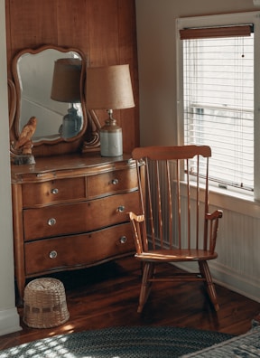 A cozy corner of a room features a wooden dresser with a mirror on top, a classic wooden chair, a table lamp with a beige shade, and a decorative bird figurine. The light from the window with blinds softly illuminates the wooden furniture and creates a warm atmosphere.