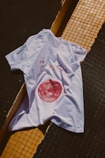 A white t-shirt is laid over a tiled surface that consists of black and yellow tiles. The t-shirt has a graphic design featuring a red circular image resembling a celestial body, along with some text and arrows.