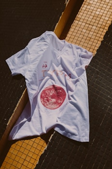 A white t-shirt is laid over a tiled surface that consists of black and yellow tiles. The t-shirt has a graphic design featuring a red circular image resembling a celestial body, along with some text and arrows.