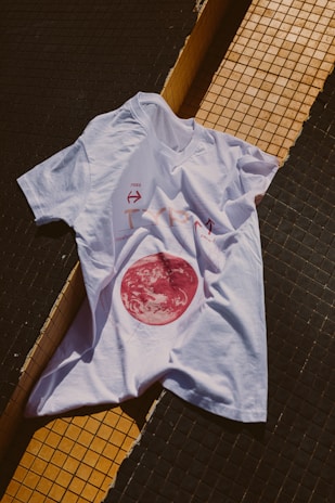 A white t-shirt is laid over a tiled surface that consists of black and yellow tiles. The t-shirt has a graphic design featuring a red circular image resembling a celestial body, along with some text and arrows.