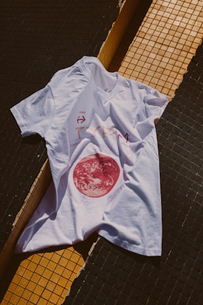 A white t-shirt is laid over a tiled surface that consists of black and yellow tiles. The t-shirt has a graphic design featuring a red circular image resembling a celestial body, along with some text and arrows.