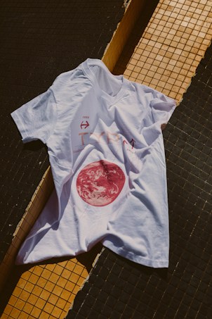 A white t-shirt is laid over a tiled surface that consists of black and yellow tiles. The t-shirt has a graphic design featuring a red circular image resembling a celestial body, along with some text and arrows.