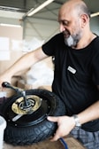 A person is working with a tire and holding it with one hand while using a tool with the other hand. They are in a workshop environment filled with boxes and work-related materials.