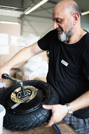 A person is working with a tire and holding it with one hand while using a tool with the other hand. They are in a workshop environment filled with boxes and work-related materials.