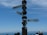 A signpost displays three directional arrows pointing towards Sydney, Berlin, and London with respective distances. The background features a clear blue sky, and the horizon showcases faint outlines of distant mountains. Several people are partially visible at the bottom of the image, indicating a viewpoint or tourist location.
