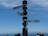 A signpost displays three directional arrows pointing towards Sydney, Berlin, and London with respective distances. The background features a clear blue sky, and the horizon showcases faint outlines of distant mountains. Several people are partially visible at the bottom of the image, indicating a viewpoint or tourist location.