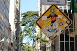 A street sign is heavily covered with various colorful stickers and decals, some featuring text and graphic designs. The background reveals a cityscape with tall buildings and a leafy tree under a clear sky.