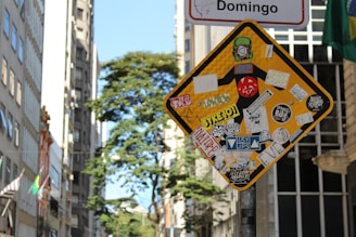 A street sign is heavily covered with various colorful stickers and decals, some featuring text and graphic designs. The background reveals a cityscape with tall buildings and a leafy tree under a clear sky.