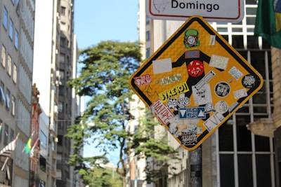 A street sign is heavily covered with various colorful stickers and decals, some featuring text and graphic designs. The background reveals a cityscape with tall buildings and a leafy tree under a clear sky.
