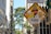 A street sign is heavily covered with various colorful stickers and decals, some featuring text and graphic designs. The background reveals a cityscape with tall buildings and a leafy tree under a clear sky.