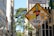 A street sign is heavily covered with various colorful stickers and decals, some featuring text and graphic designs. The background reveals a cityscape with tall buildings and a leafy tree under a clear sky.