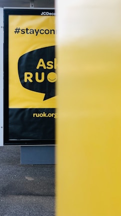 A close-up view of a public advertisement with a yellow background, partially obscured by a yellow object in the foreground. The words 'Ask R U OK?' are visible inside a speech bubble design. Above that, '#stayconnected' is written, and at the bottom, 'ruok.org' is printed.