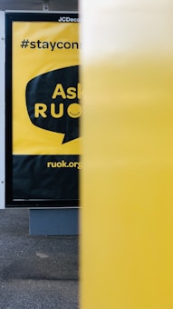 A close-up view of a public advertisement with a yellow background, partially obscured by a yellow object in the foreground. The words 'Ask R U OK?' are visible inside a speech bubble design. Above that, '#stayconnected' is written, and at the bottom, 'ruok.org' is printed.