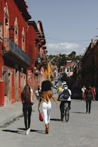 A vibrant street scene features colorful buildings with ornate architectural details, lining a cobblestone street bustling with people. Two women walk side by side, while a cyclist and other pedestrians navigate the lively atmosphere. The sky is clear with scattered clouds, adding to the dynamic urban environment.