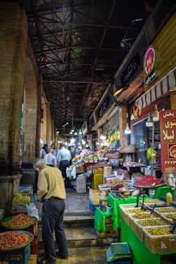 A bustling indoor market aisle lined with various stalls offering fresh produce. The left side features a man selecting items from a basket of fruits or vegetables. The right side displays an assortment of goods including olives, nuts, and spices. Overhead, a high, metal beam ceiling gives an industrial feel, with hanging lights illuminating the corridor. Several people are seen further down the aisle engaging in shopping and browsing.
