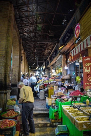 A bustling indoor market aisle lined with various stalls offering fresh produce. The left side features a man selecting items from a basket of fruits or vegetables. The right side displays an assortment of goods including olives, nuts, and spices. Overhead, a high, metal beam ceiling gives an industrial feel, with hanging lights illuminating the corridor. Several people are seen further down the aisle engaging in shopping and browsing.