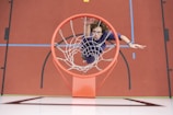 A person in sports attire is captured from above reaching towards a basketball hoop, appearing to be in the act of making a dunk. The court features vibrant colors including red, blue, and black markings.