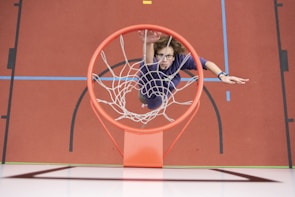 A person in sports attire is captured from above reaching towards a basketball hoop, appearing to be in the act of making a dunk. The court features vibrant colors including red, blue, and black markings.